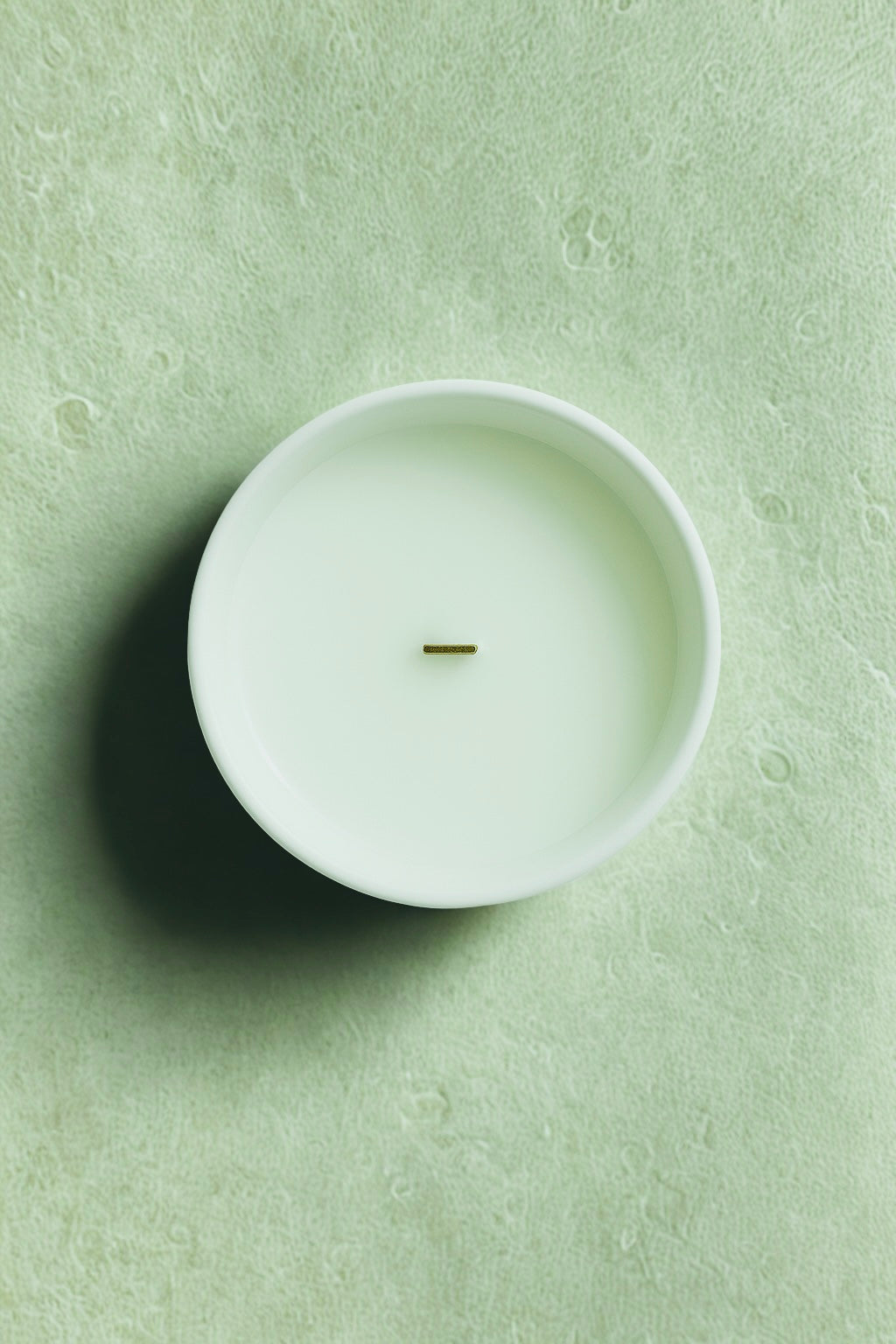 White candle in a white container on a light green textured surface“Minimalist luxury winter candle UK by Aurum Blanc, hand-poured Brume Alpine premium scented candle with alpine forest fragrance.”