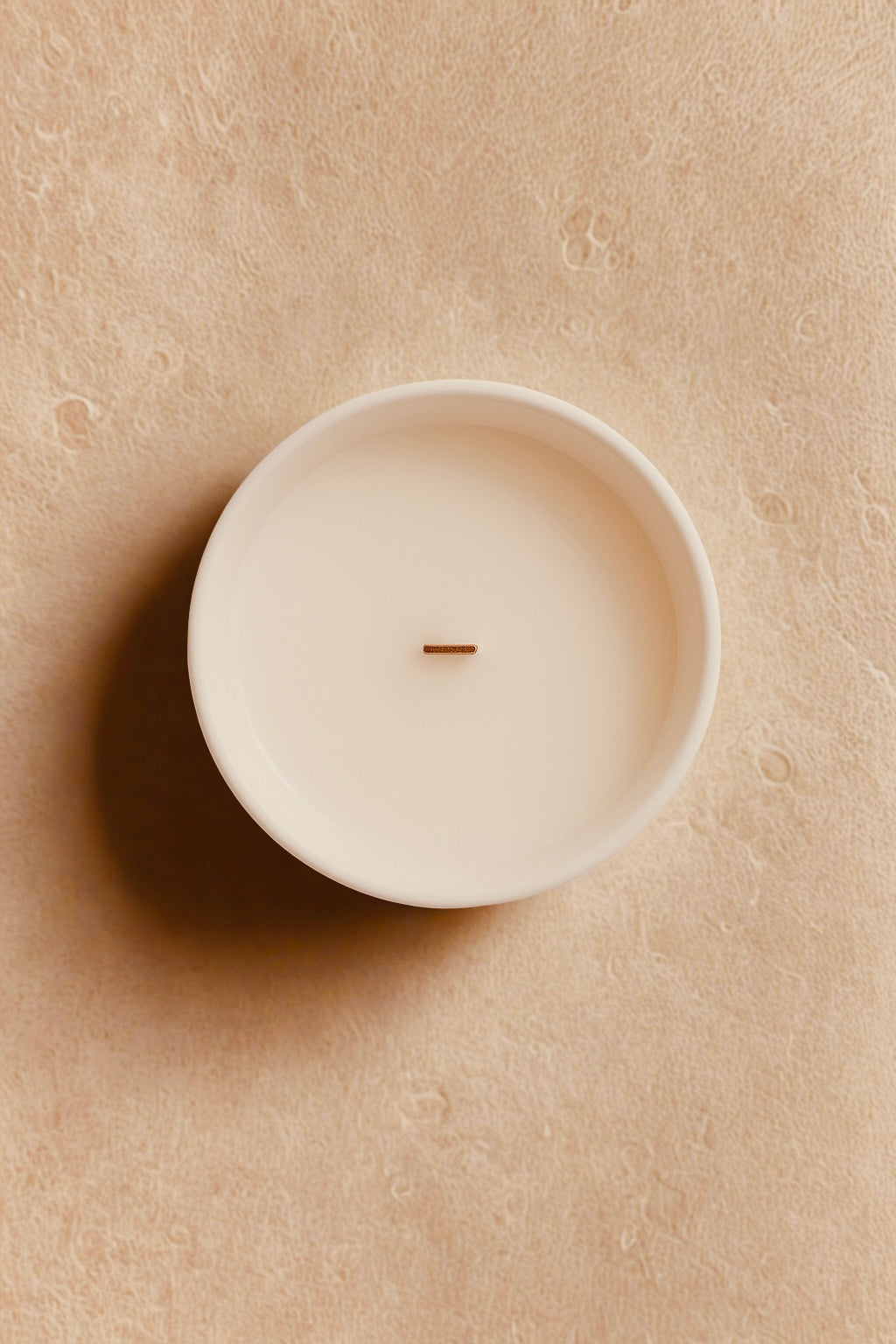 White candle in a ceramic holder on a beige textured surface“Minimalist luxury winter candle UK by Aurum Blanc, hand-poured Pommes Épicées premium scented candle with apple and ginger fragrance.”